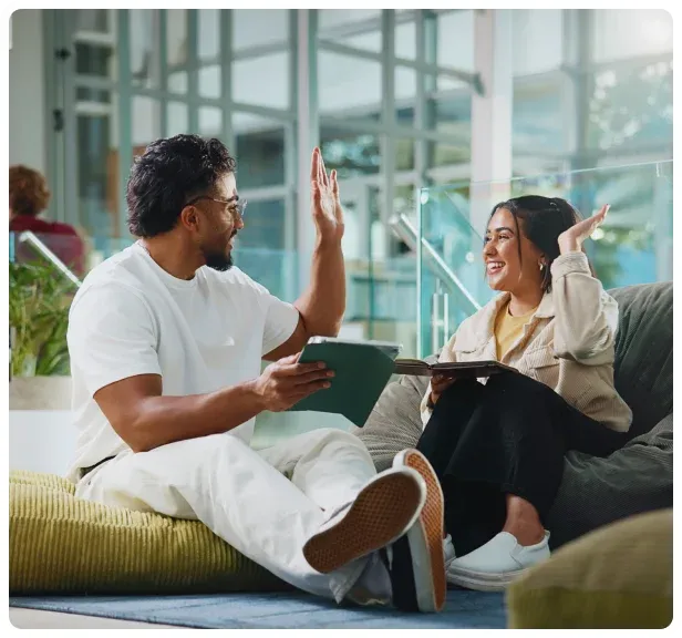 Two coworkers sitting in a bright workplace lounge and celebrating progress together.